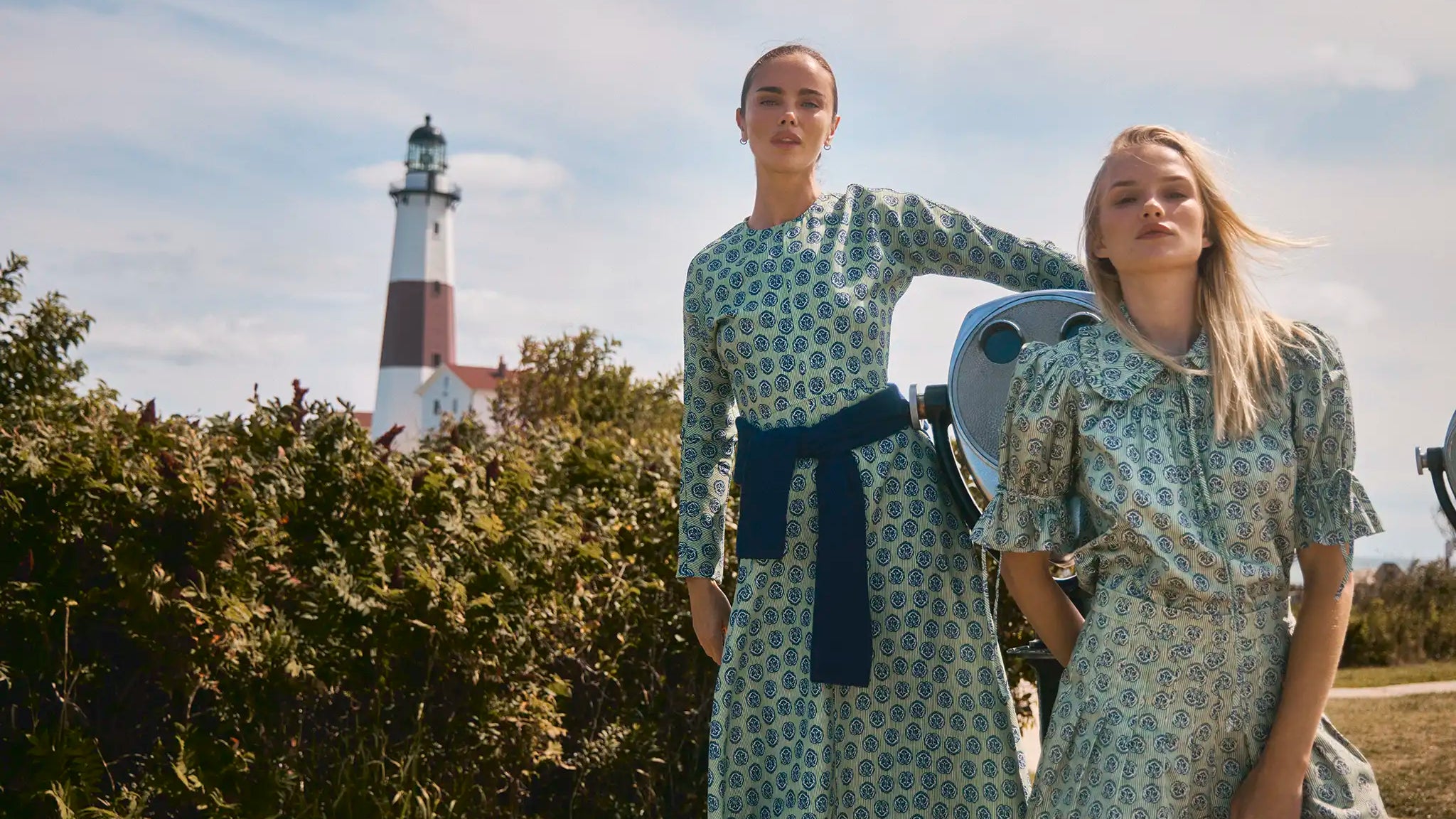 Two girls - one in a green longsleeve dress with navy accents other girl in same print but matching top and short in front of lighthouse