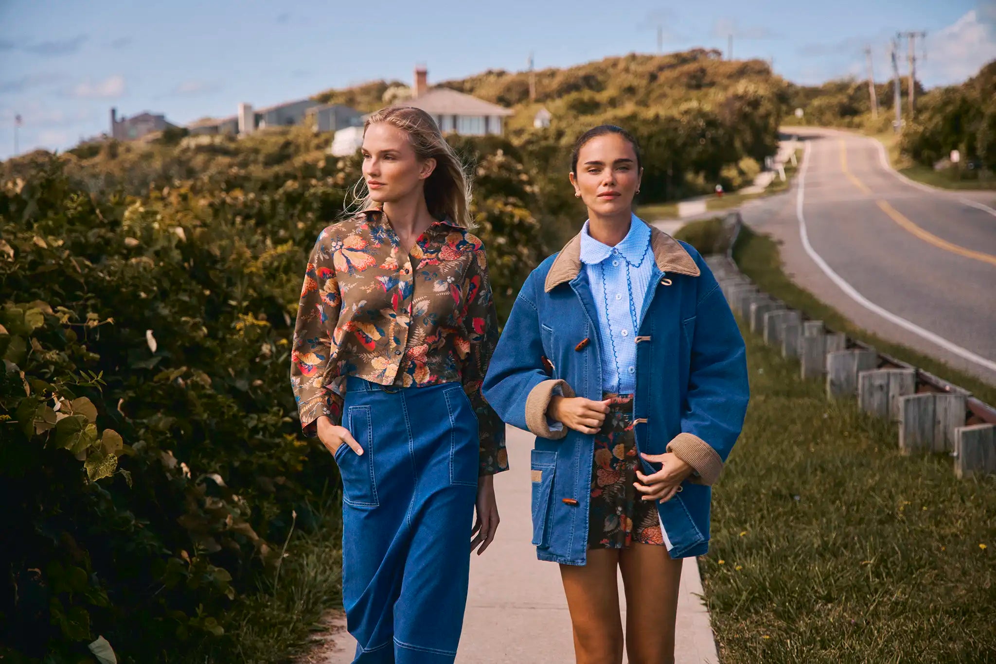 Two women walking along a road with greenery and houses in the background