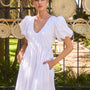 The Augusta Dress, white cotton dress with ruched V neckline, in front of a fence in the sun.