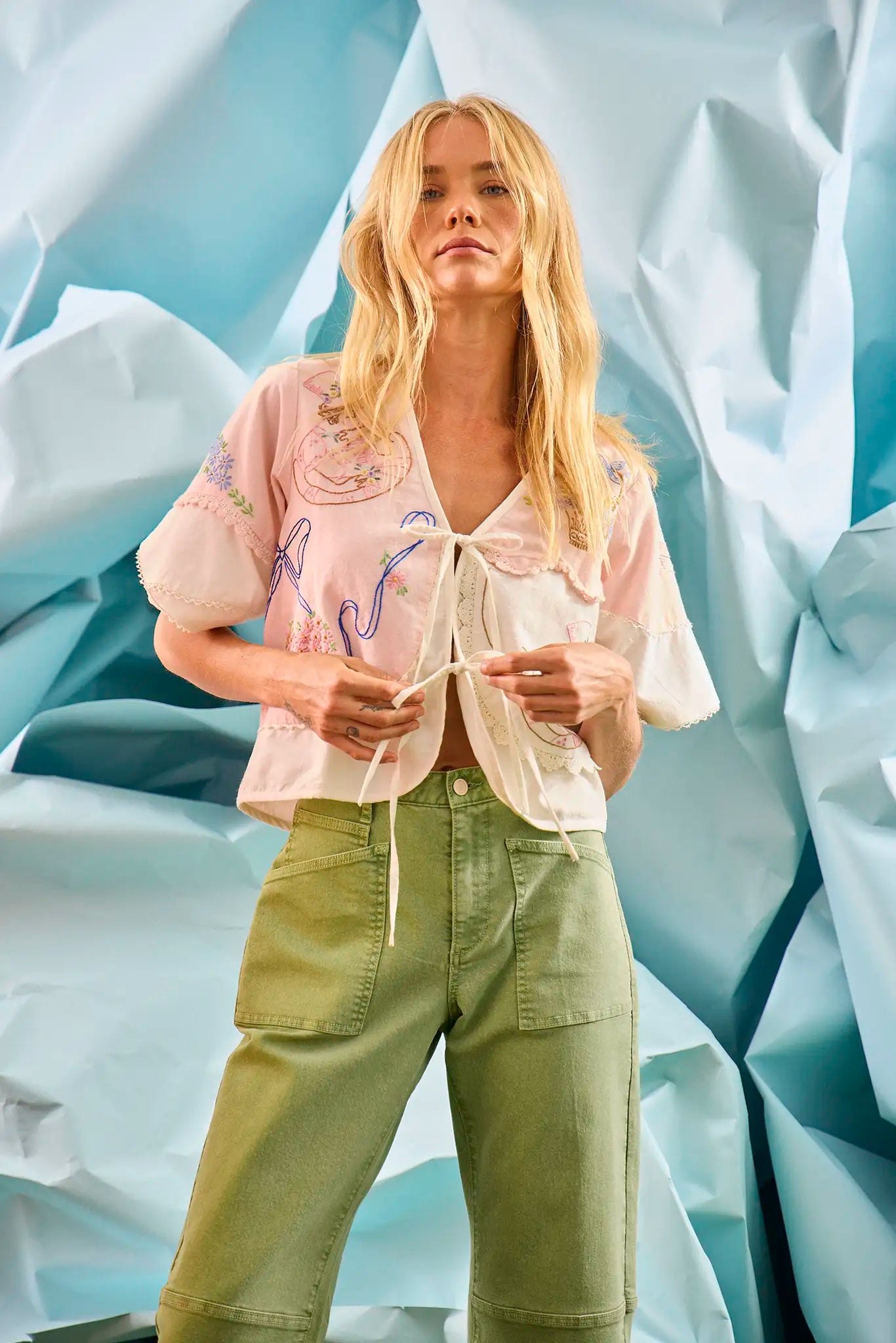The Bryce Top in Lazy Daisy paired with green Mallory Jeans in front of blue wall.