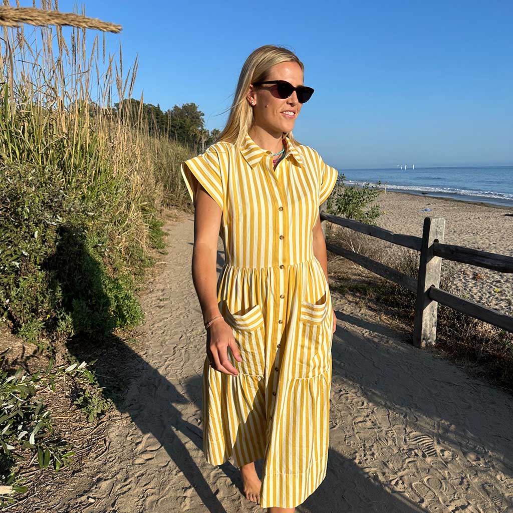 Blond woman wearing a yellow and white stripes jumper walking on a beach and smiling